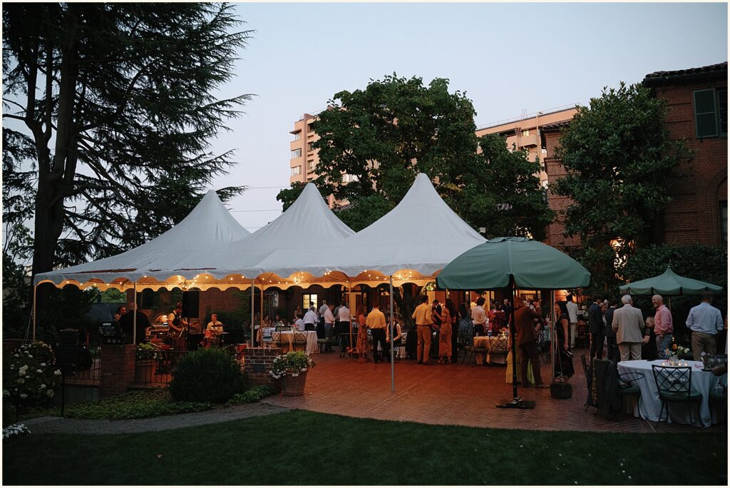 Wedding guests gather under a white tent in the evening.